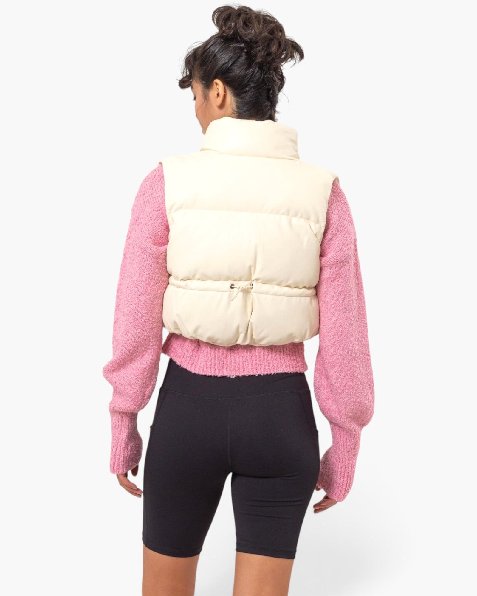 A model shown from the back wearing a cream puffer vest over a pink textured knit sweater and black bike shorts, against a plain white studio background.