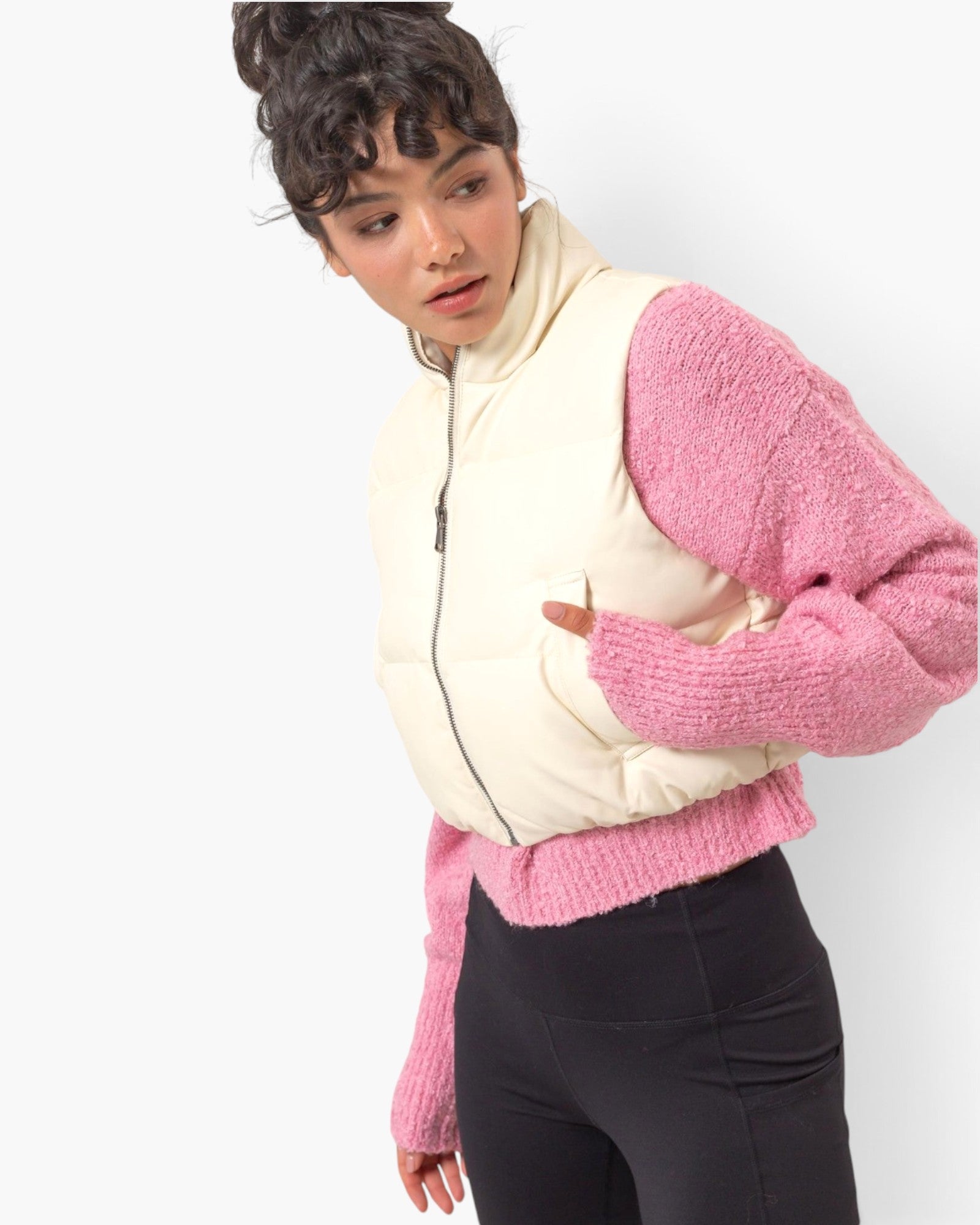A model wearing a cream-colored puffer vest with pink knit sleeves and a pink ribbed hem, paired with black pants, against a plain white background.