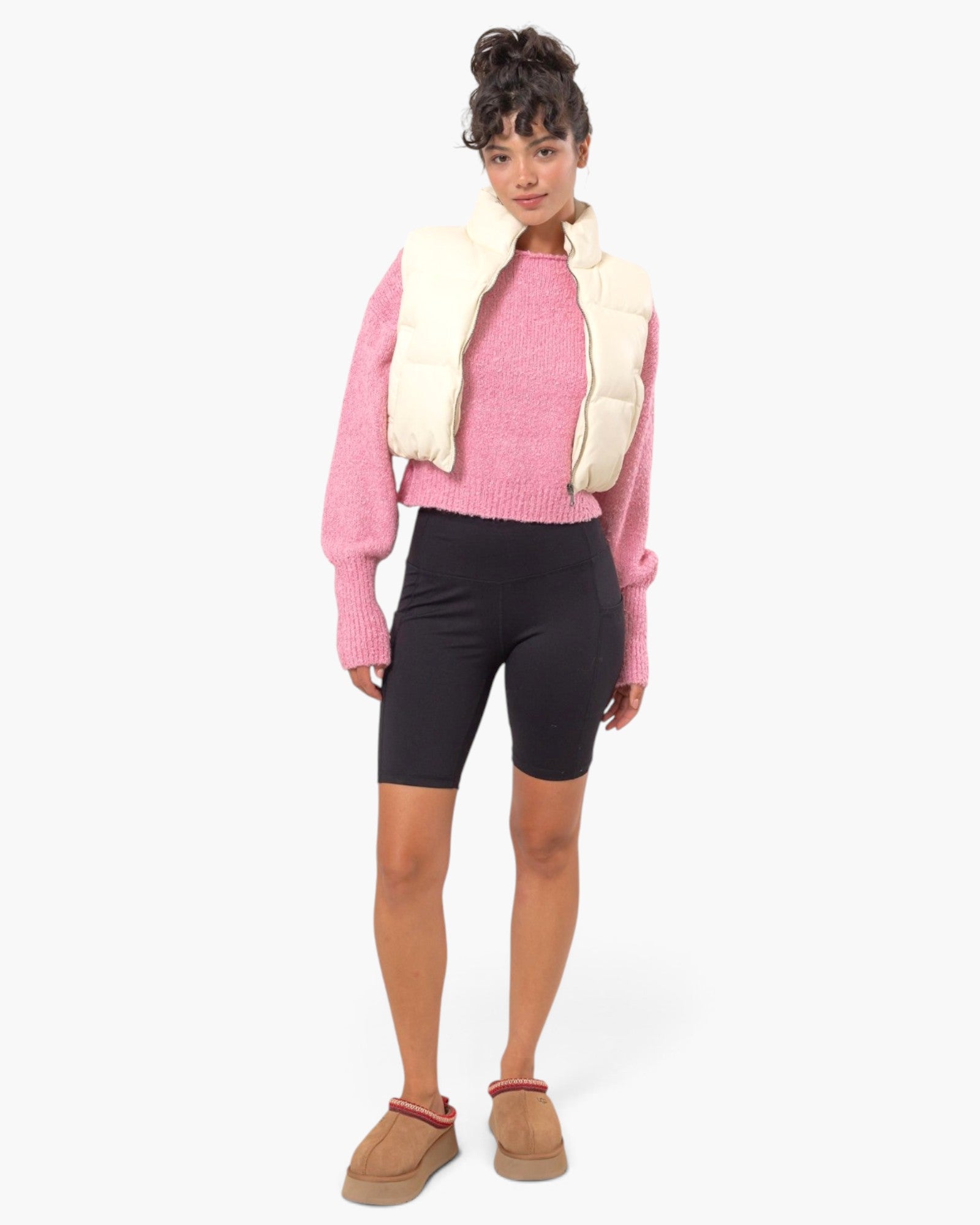 A model stands against a light gray studio background wearing a pink knit sweater, a cream puffer vest draped over the shoulders, black biker shorts, and tan slip-on mule shoes.