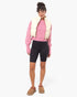 A model stands against a light gray studio background wearing a pink knit sweater, a cream puffer vest draped over the shoulders, black biker shorts, and tan slip-on mule shoes.
