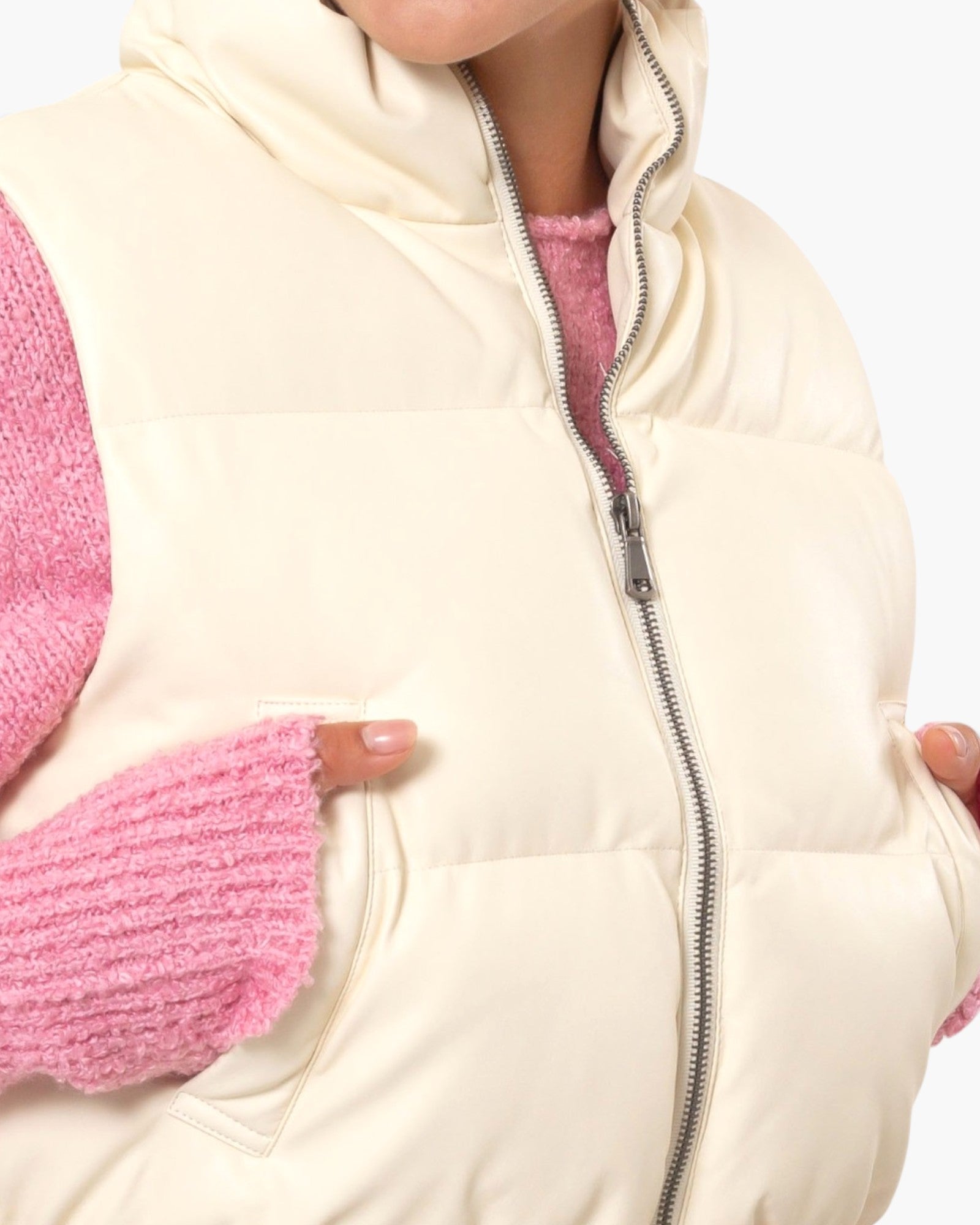 Close-up of a cream puffer vest worn over a pink knit sweater, showing the front zipper, high collar, and side pockets with hands in the pockets.