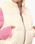 Close-up of a cream puffer vest worn over a pink knit sweater, showing the front zipper, high collar, and side pockets with hands in the pockets.