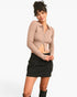A woman modeling a beige ribbed long-sleeve crop top with a front zipper and a black denim mini skirt, standing against a plain white studio background.