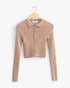 A beige ribbed knit long-sleeve crop top with a collar and front zipper, hanging on a wooden hanger against a plain white background.