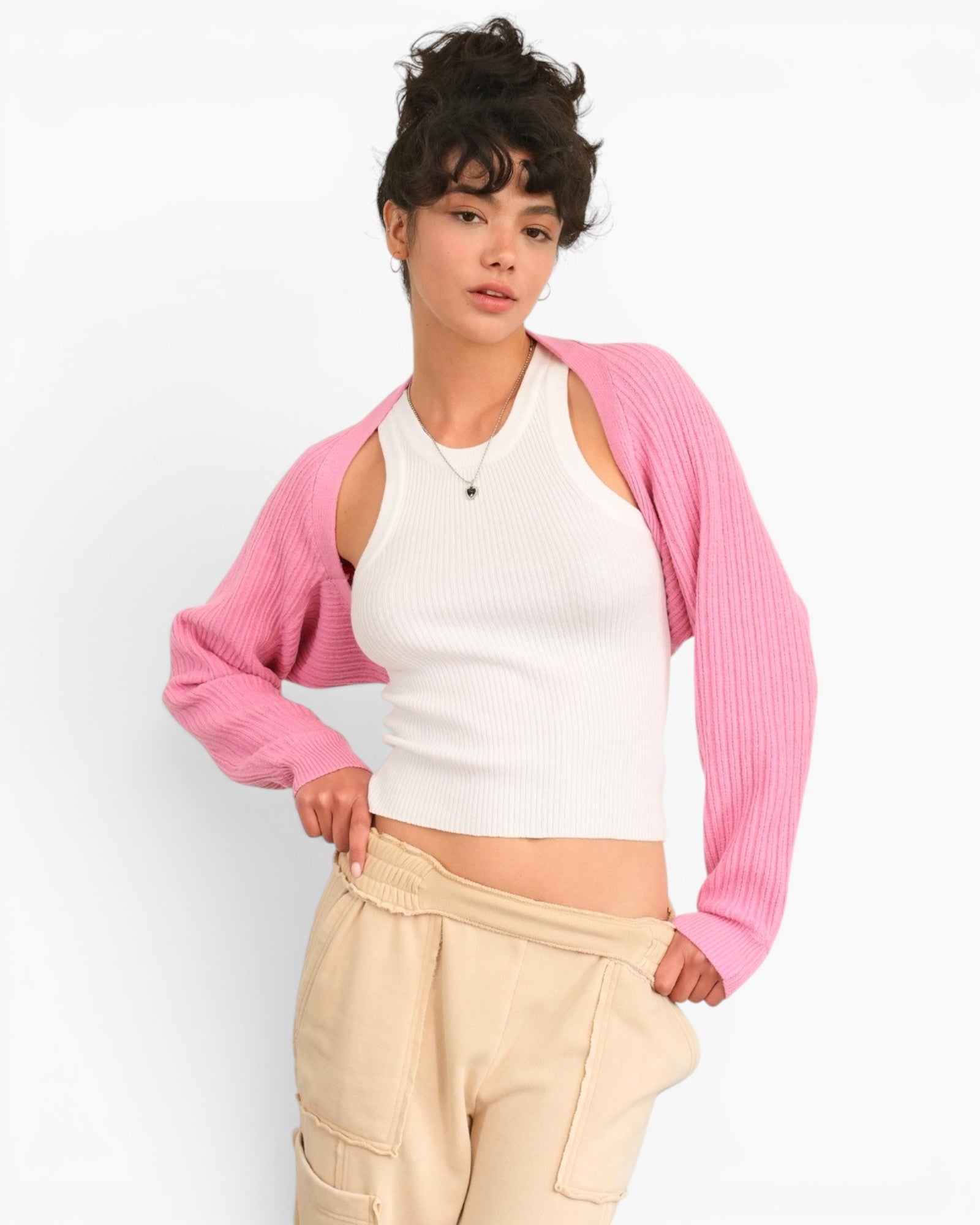 A model wearing a pink ribbed knit shrug cardigan over a white ribbed tank top and beige cargo pants, posed against a clean white studio background.