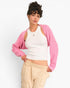 A model wearing a pink ribbed knit shrug cardigan over a white ribbed tank top and beige cargo pants, posed against a clean white studio background.