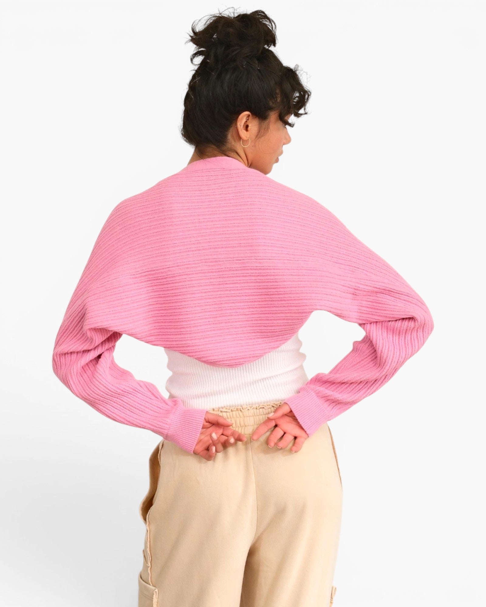 A woman shown from the back wearing a pink ribbed knit cropped sweater with long sleeves, a white top, and beige pants against a plain white background.