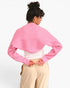 A woman shown from the back wearing a pink ribbed knit cropped sweater with long sleeves, a white top, and beige pants against a plain white background.