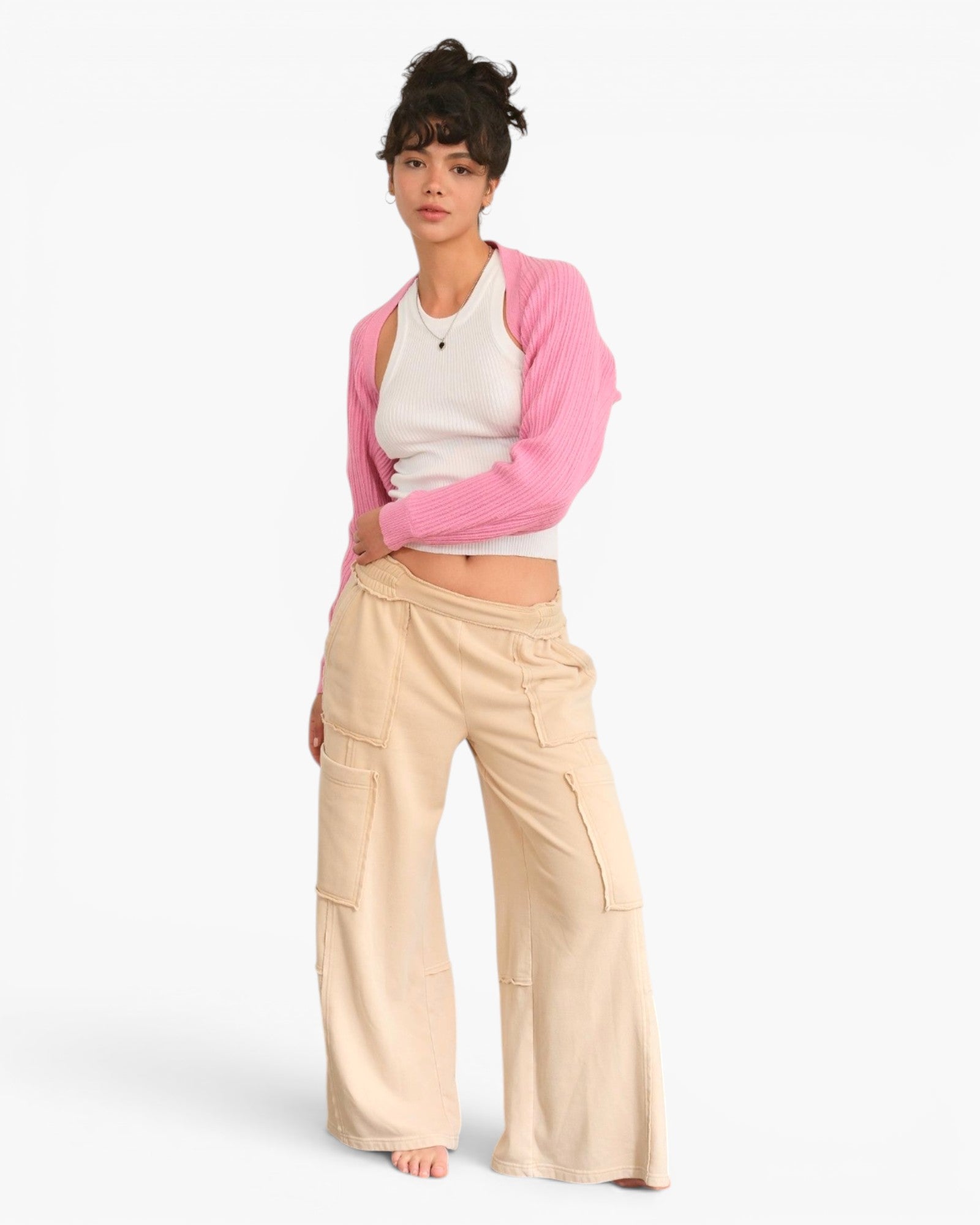A model stands against a light gray studio background wearing a pink cardigan over a white crop top and beige wide-leg cargo pants.
