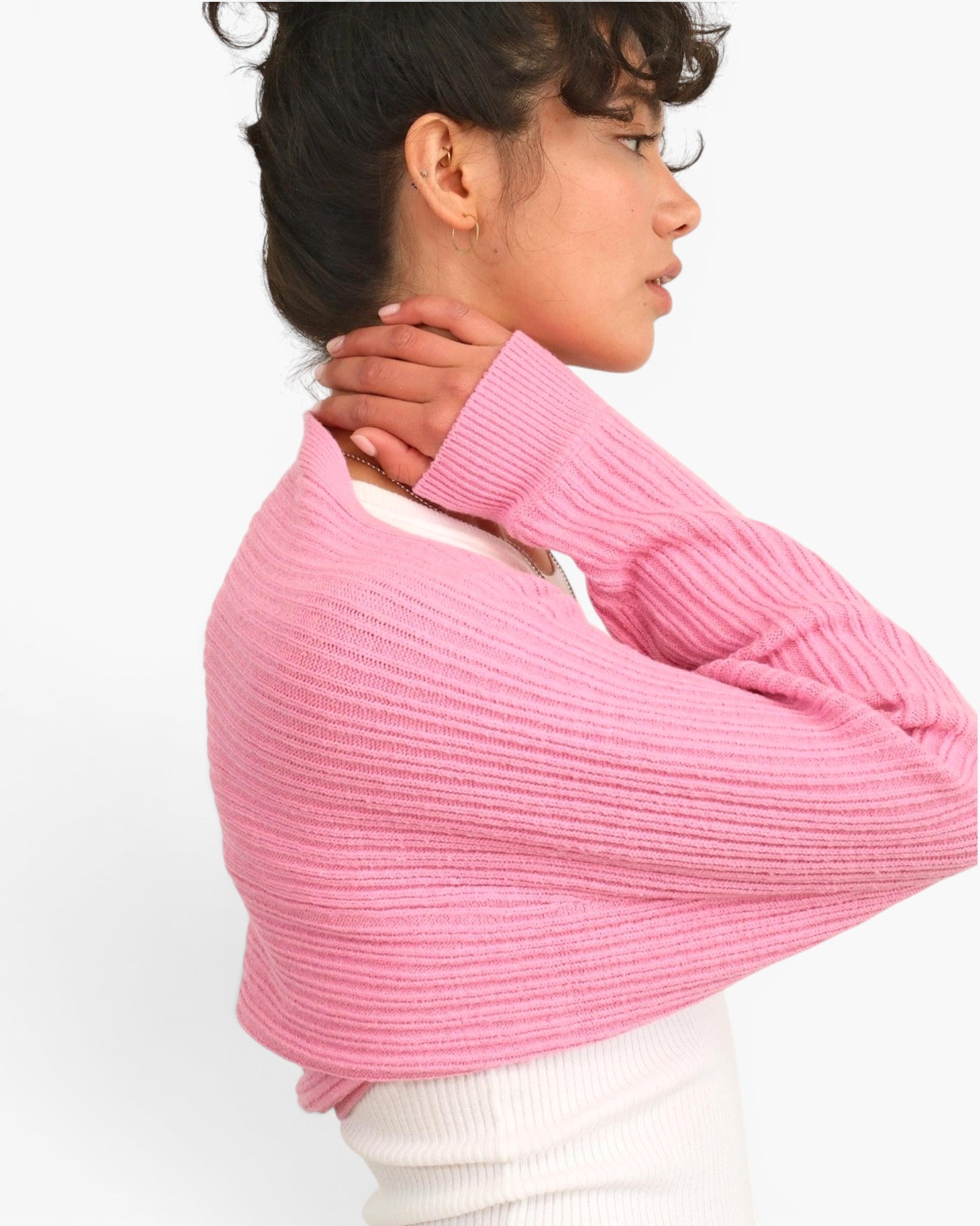A woman wearing a pink ribbed knit shrug or cropped sweater over a white top, shown in profile against a plain white background.