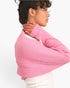 A woman wearing a pink ribbed knit shrug or cropped sweater over a white top, shown in profile against a plain white background.