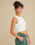 Woman wearing a white sleeveless top and green pants against a beige background