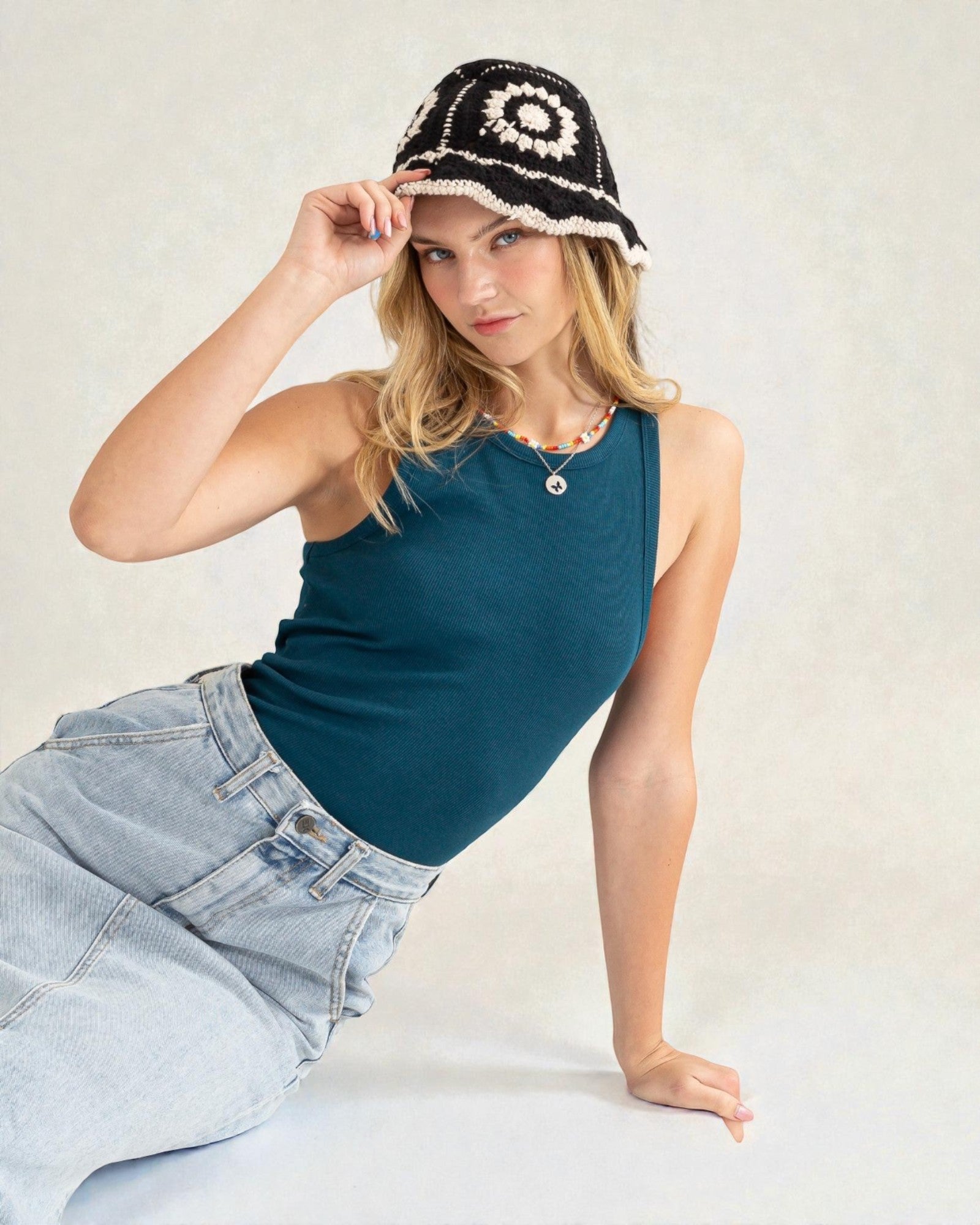 Woman wearing a teal tank top and light blue jeans, posing with a black and white patterned hat.
