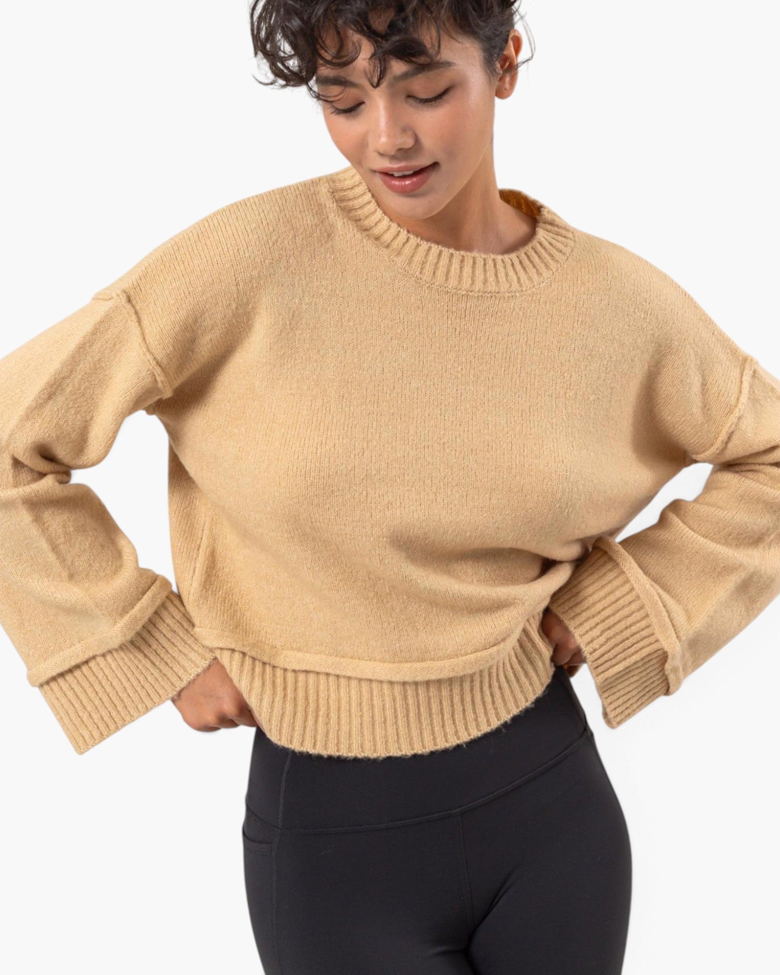 A woman wearing a cropped, long-sleeve knit sweater in a warm camel beige color, paired with black leggings, against a clean white background.