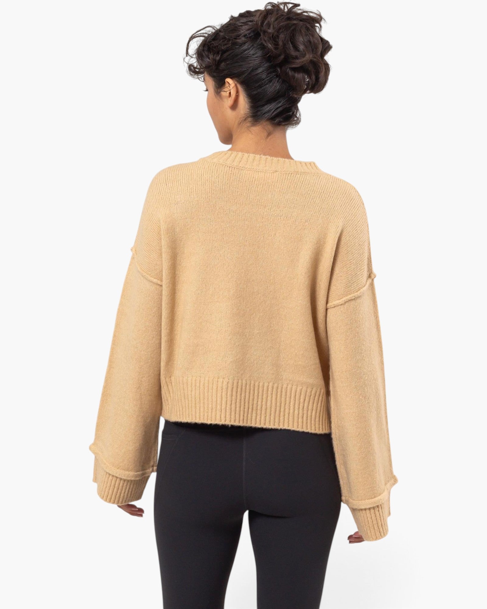 A woman shown from the back wearing a cropped, long-sleeve knit sweater in a warm camel beige color, paired with black leggings, against a plain white background.