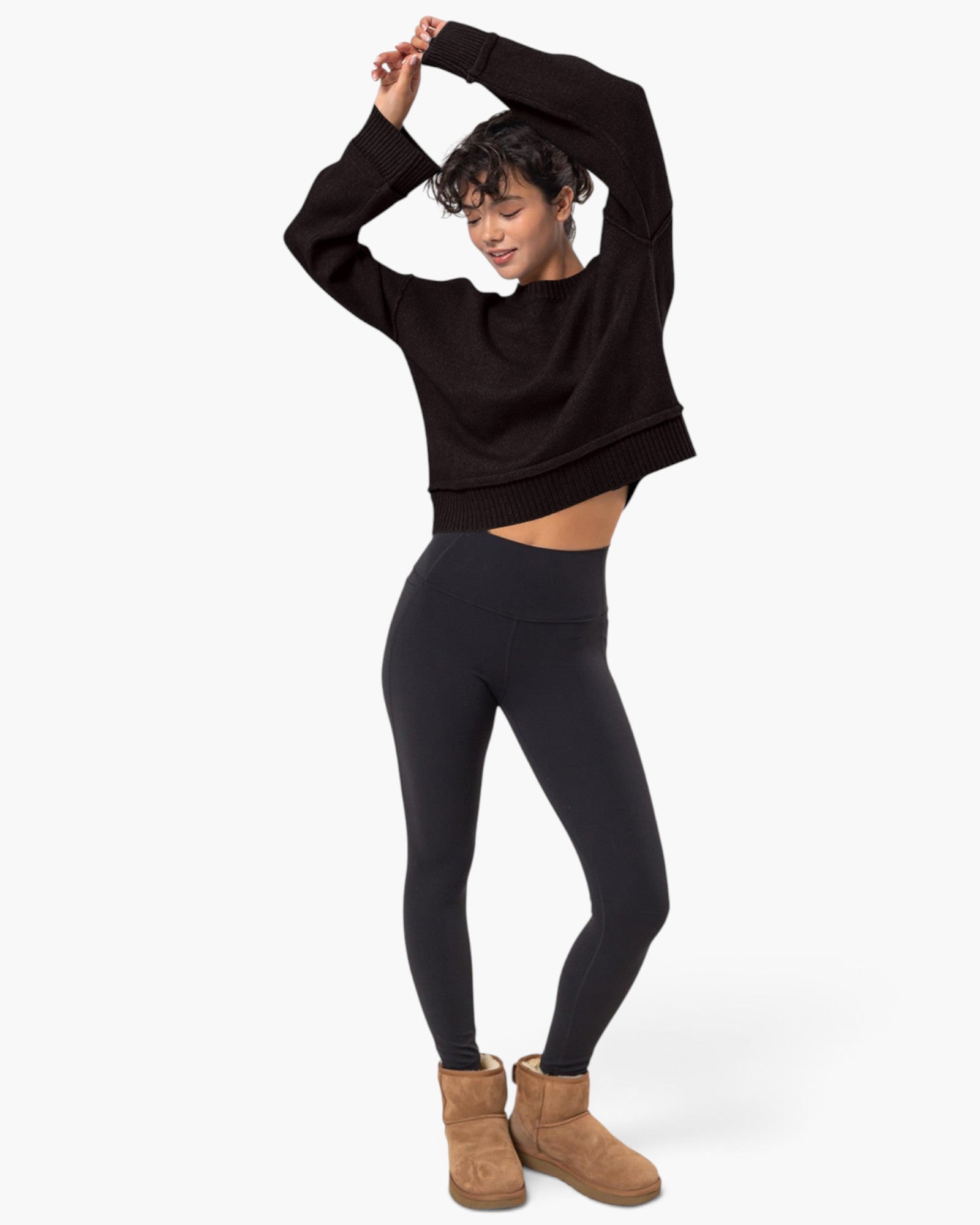 A woman modeling a black cropped long-sleeve knit top and black leggings, paired with tan ankle boots, against a plain light gray studio background.