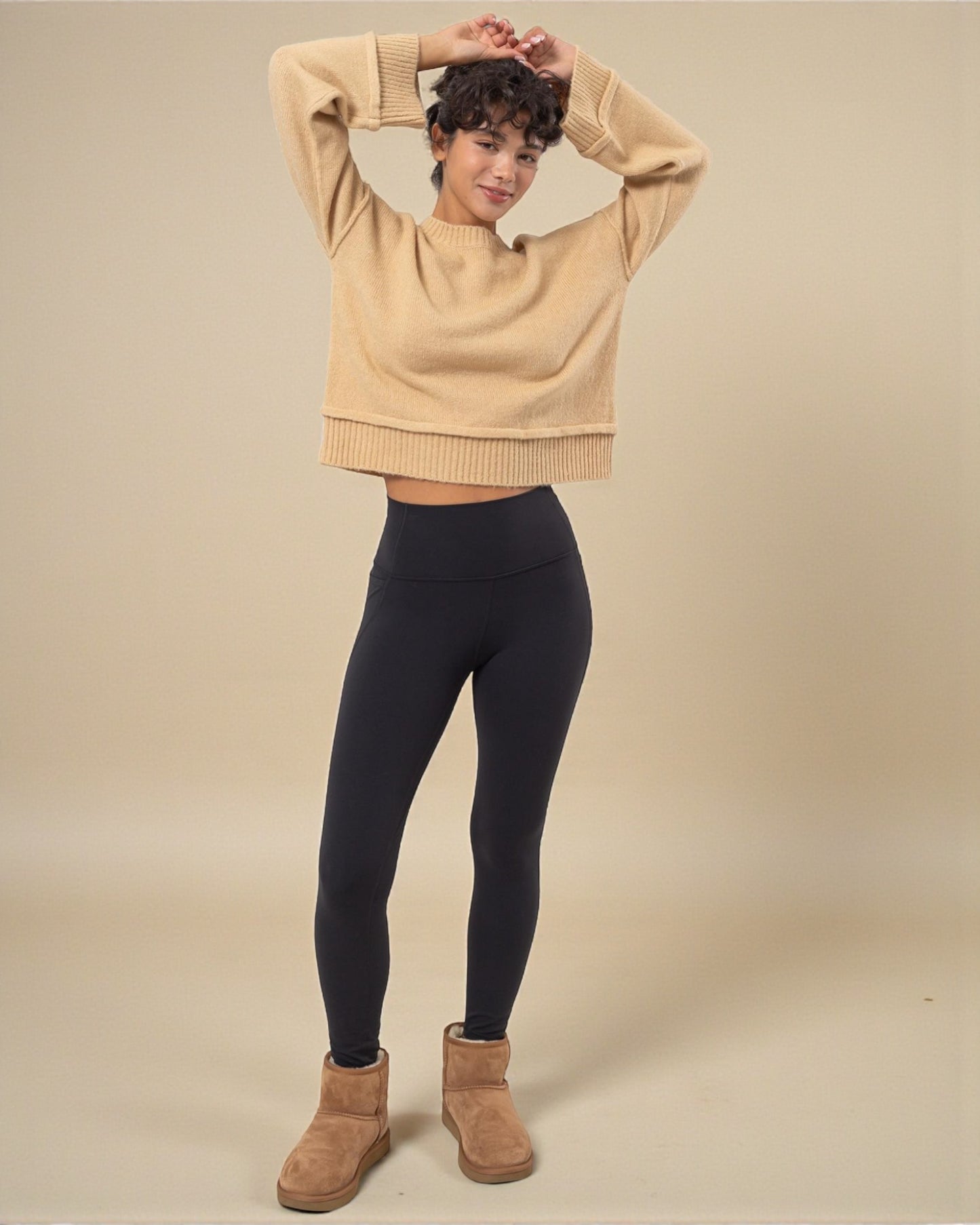 Person wearing a beige sweater, black leggings, and brown boots on a beige background