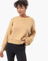 A woman wearing a cropped, long-sleeve knit sweater in a warm camel beige color, paired with black leggings, against a clean white background.