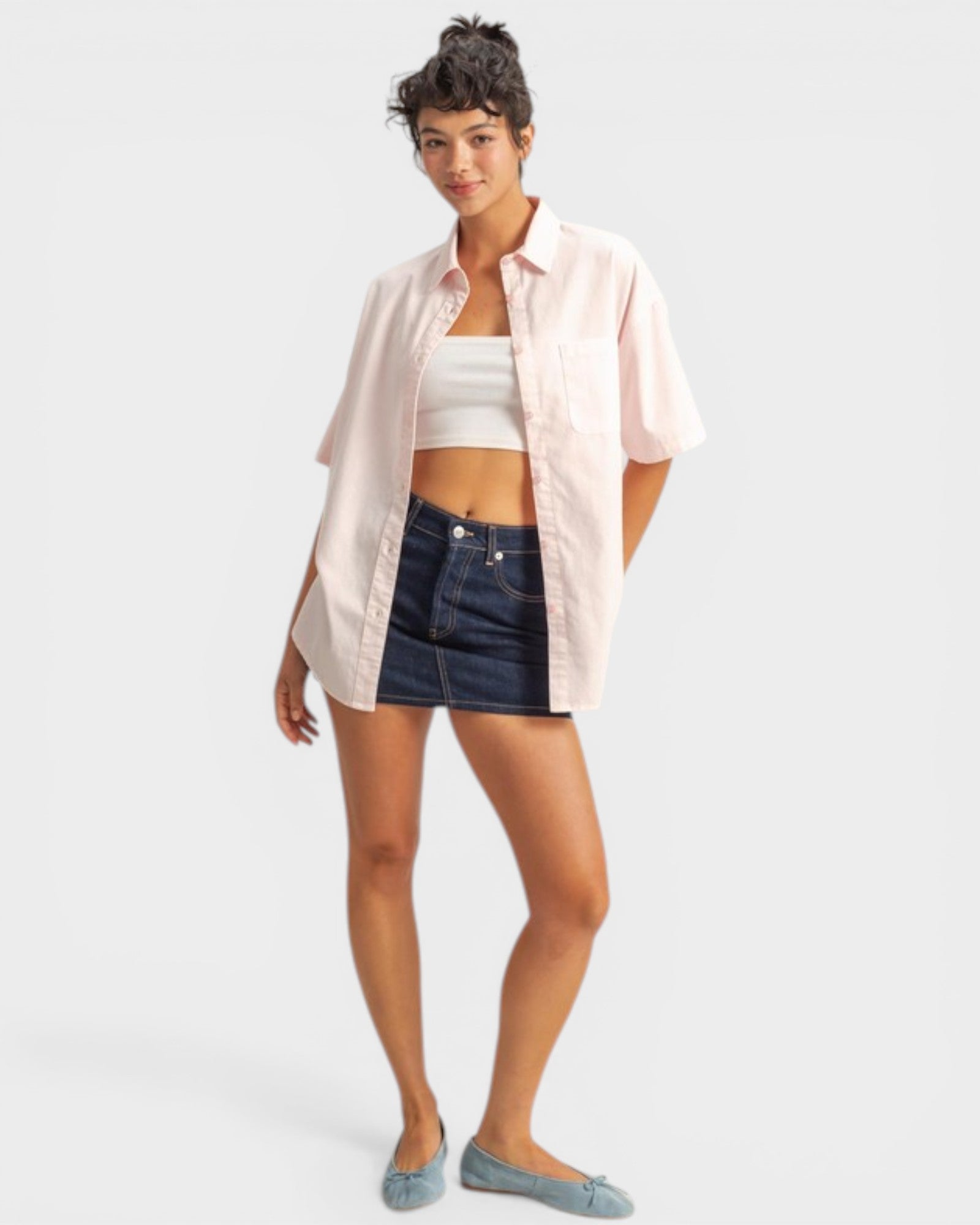 Woman wearing a light pink shirt over a white crop top and dark denim shorts on a white background