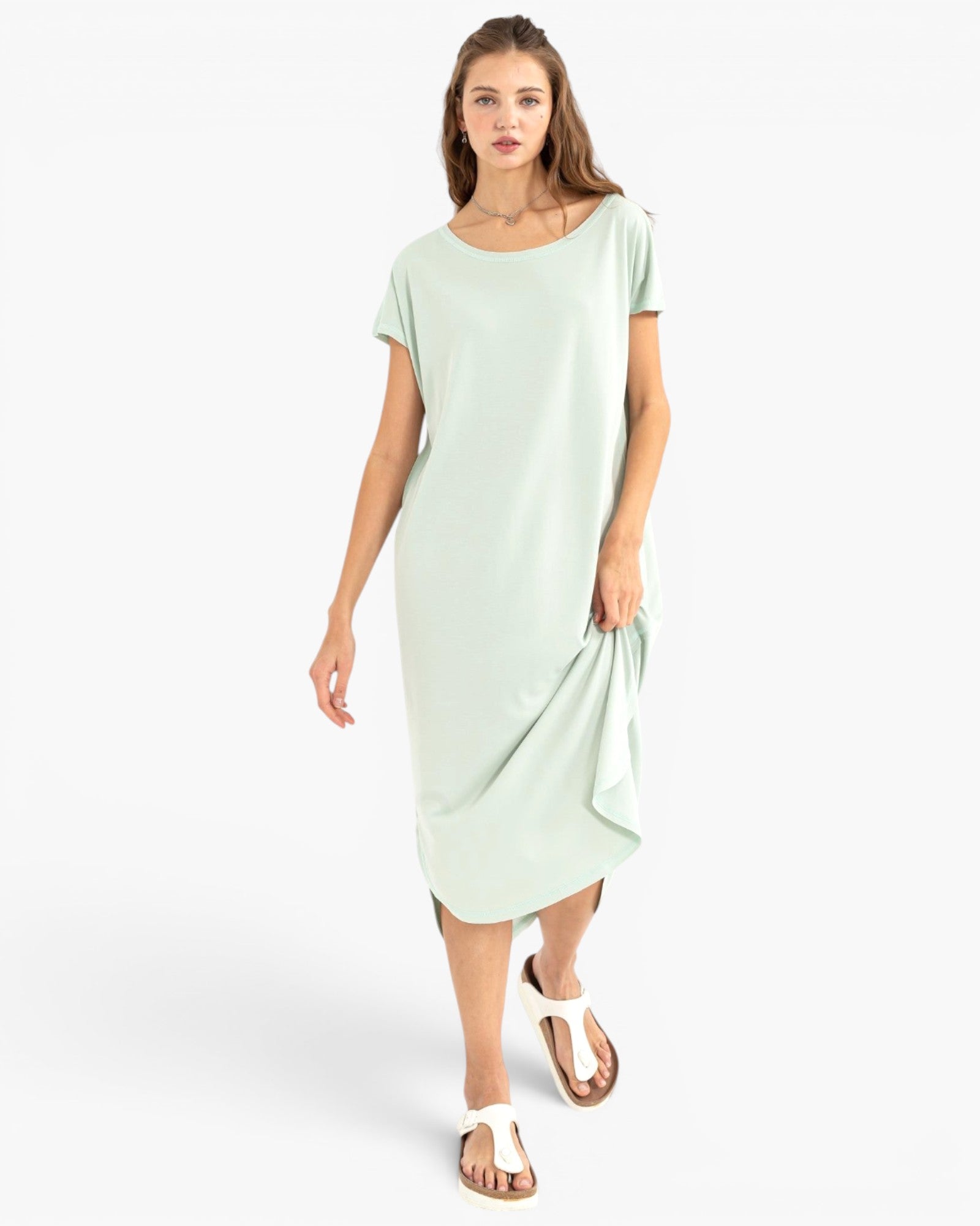 A woman modeling a light mint green short-sleeve midi dress with a draped hem, paired with white flat sandals, against a plain white studio background.