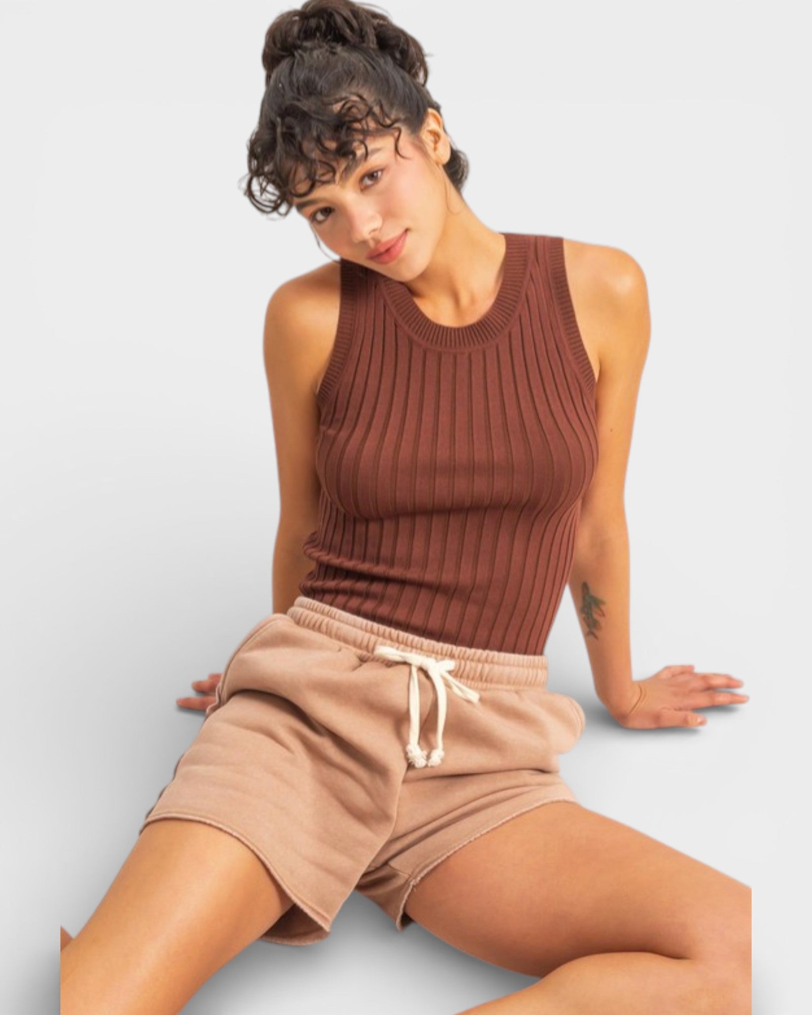 Woman wearing a brown ribbed tank top and beige shorts on a light gray background