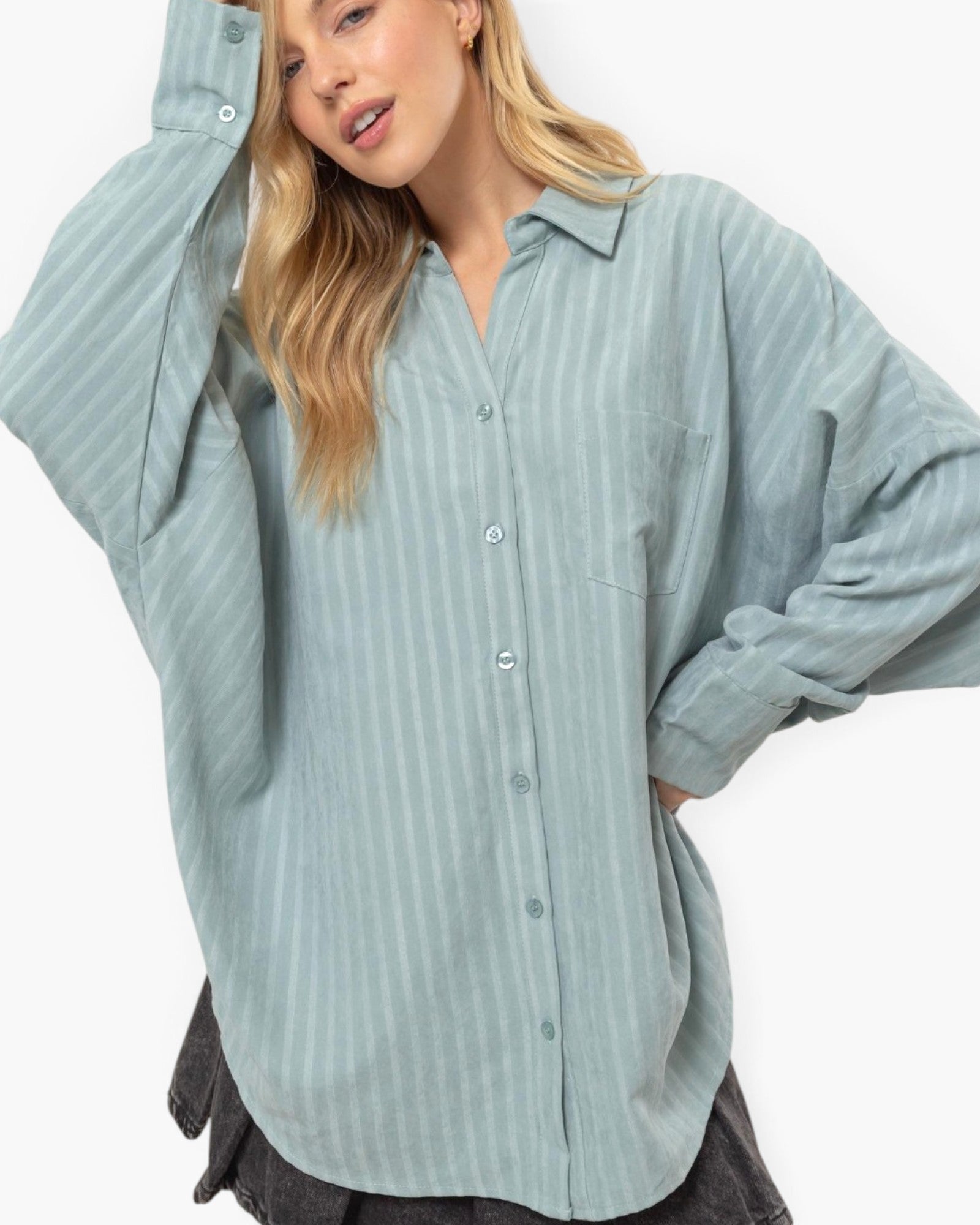 A woman wearing an oversized, long-sleeve button-up shirt in a muted sage green with thin vertical stripes, posed against a clean white background.