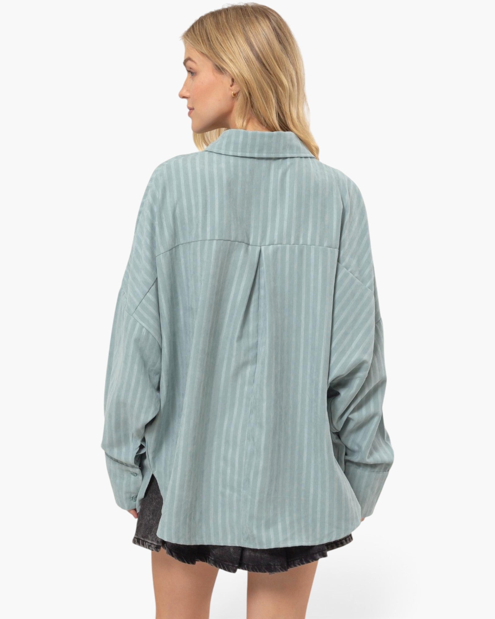 A woman shown from the back wearing an oversized sage green striped button-up shirt with dark denim shorts, set against a plain white studio background.