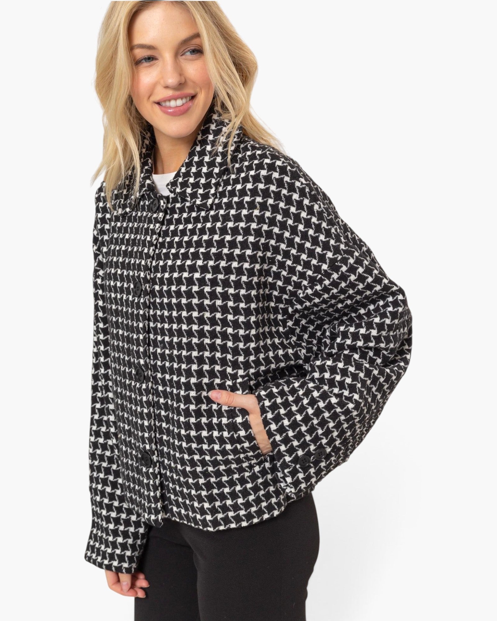 A woman wearing a black and white houndstooth patterned jacket with black pants, photographed against a clean white studio background.