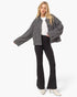 A blonde model wearing a black and white houndstooth cropped jacket, black flared pants, a white top, and white heeled boots, standing against a light gray studio background.