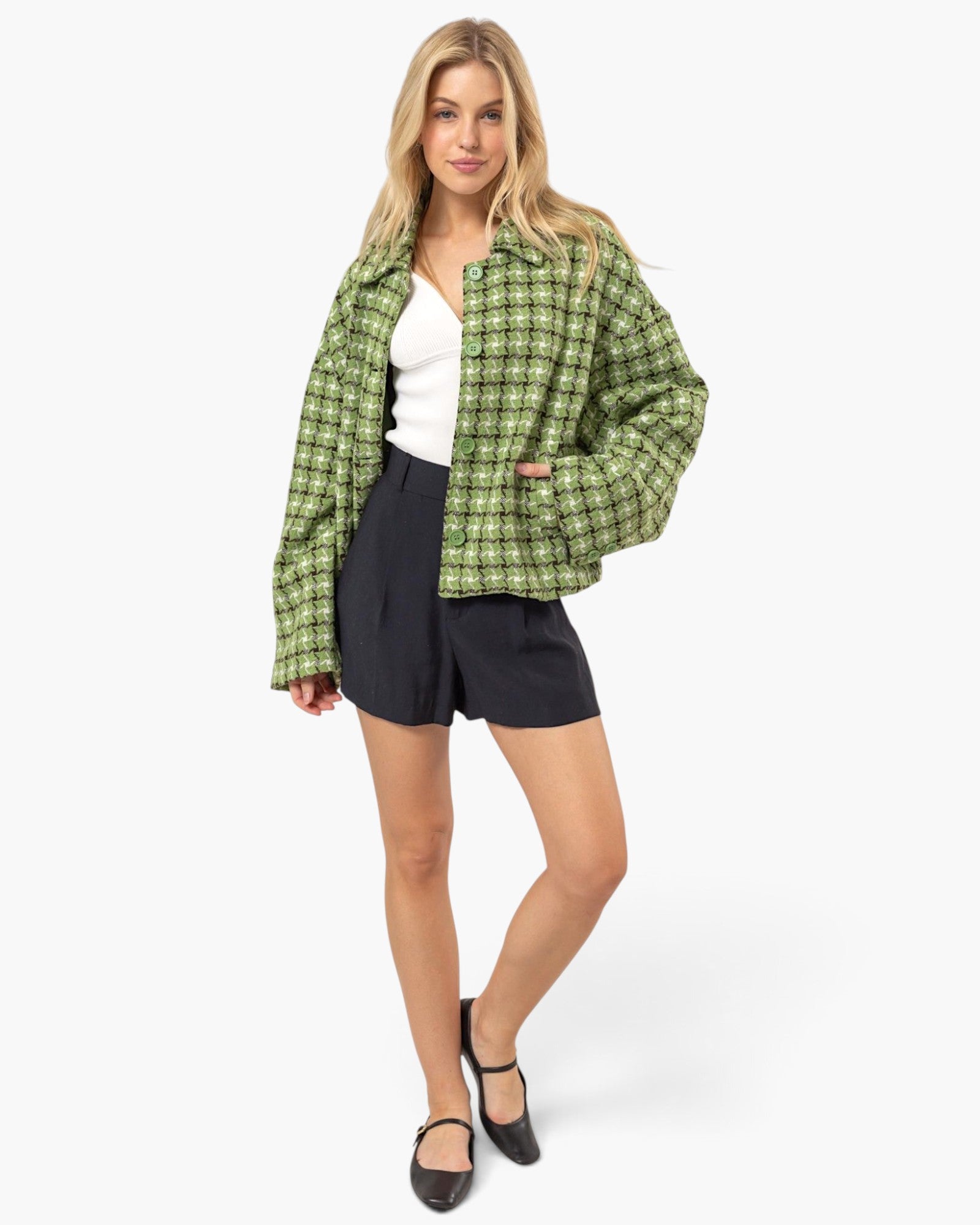 A woman modeling a green and black plaid button-up jacket over a white top and black shorts, standing against a plain white studio background.
