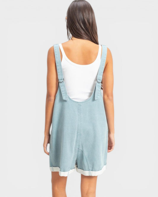 Person wearing a light blue denim overall dress on a white background