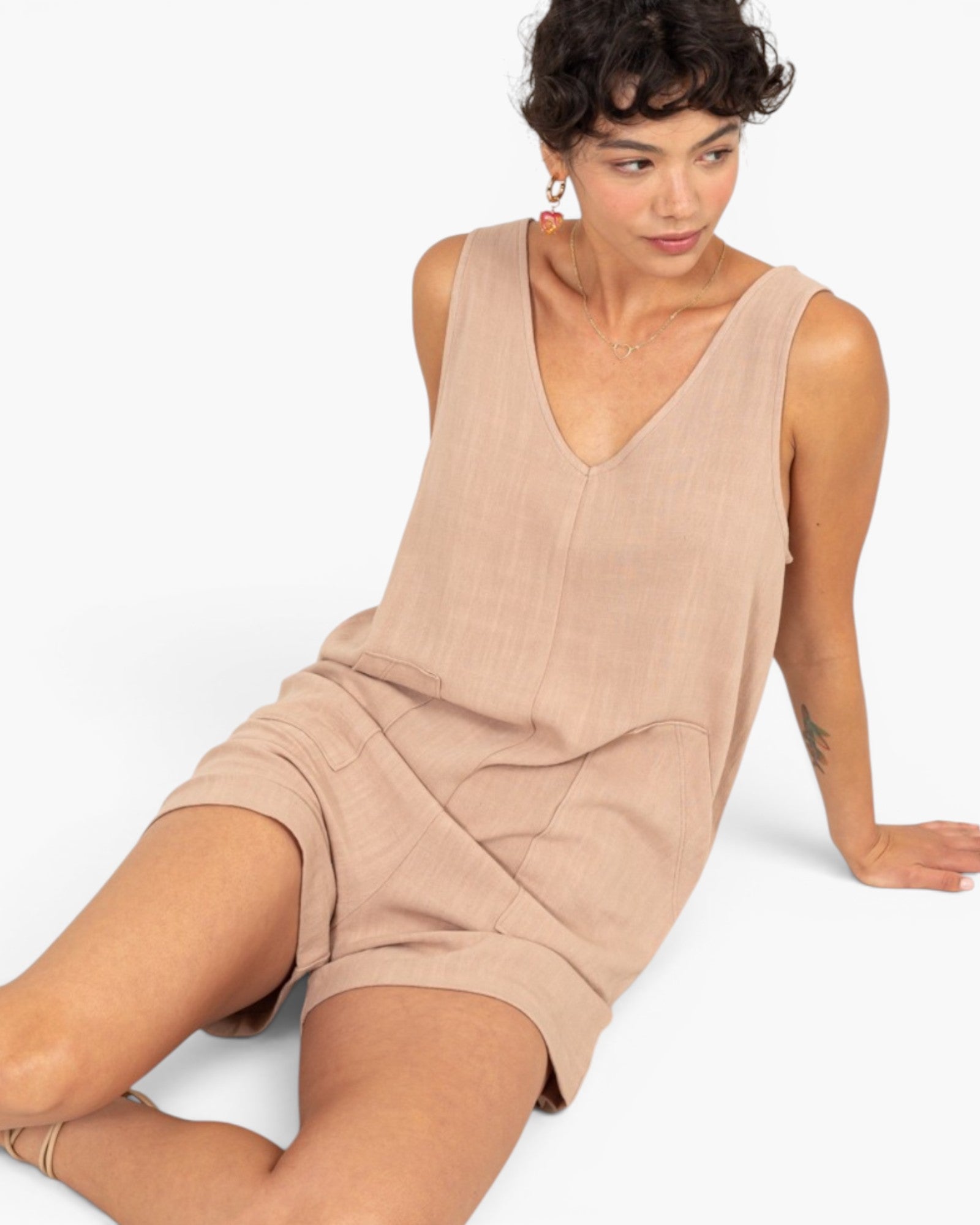 A woman wearing a sleeveless, v-neck beige linen romper, sitting against a plain white background.