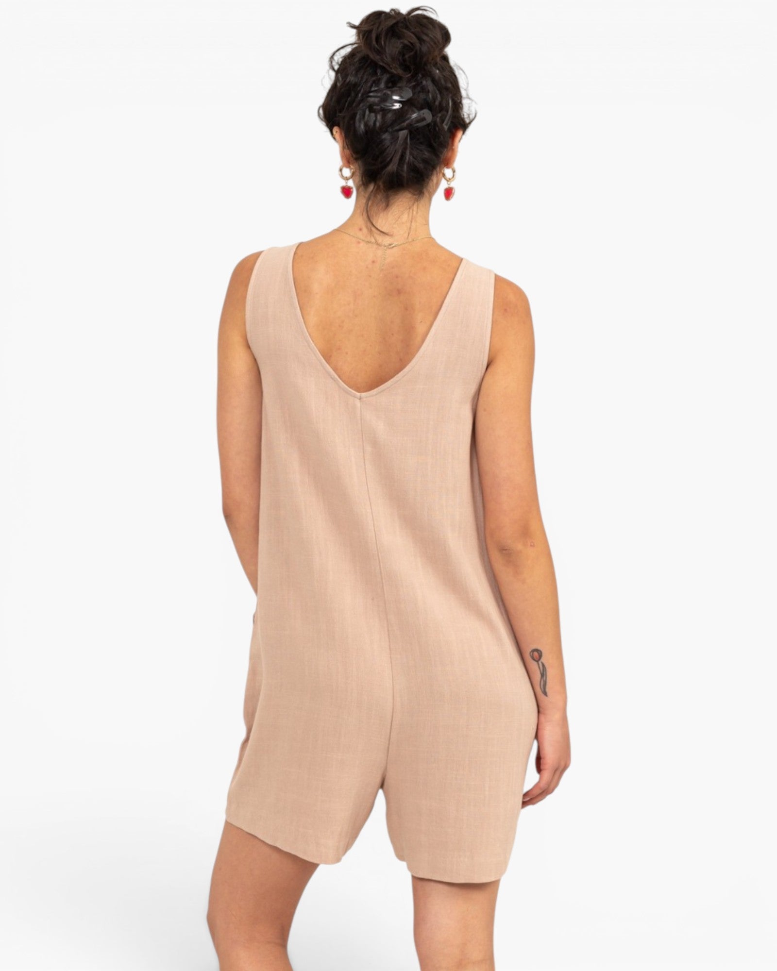 A woman shown from the back wearing a sleeveless, light beige romper with a deep V-neckline, set against a plain white background.