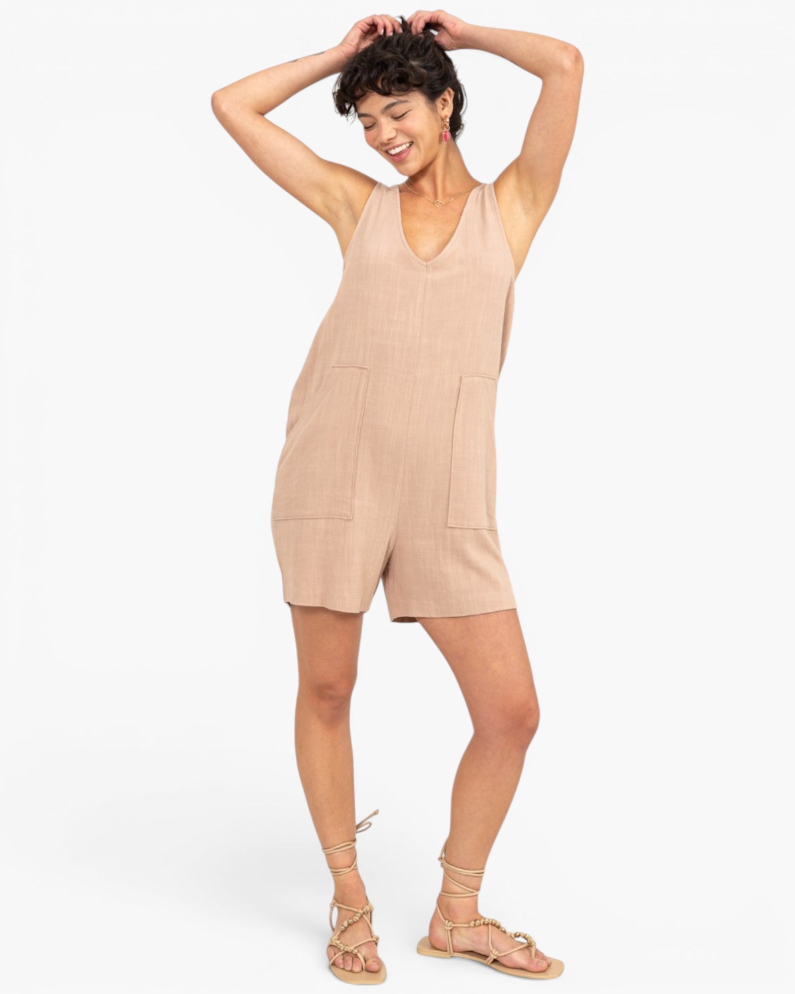 A woman modeling a sleeveless beige linen romper with a V-neck and front patch pockets, paired with beige strappy sandals, against a plain white studio background.