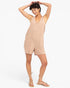 A woman modeling a sleeveless beige linen romper with a V-neck and front patch pockets, paired with beige strappy sandals, against a plain white studio background.