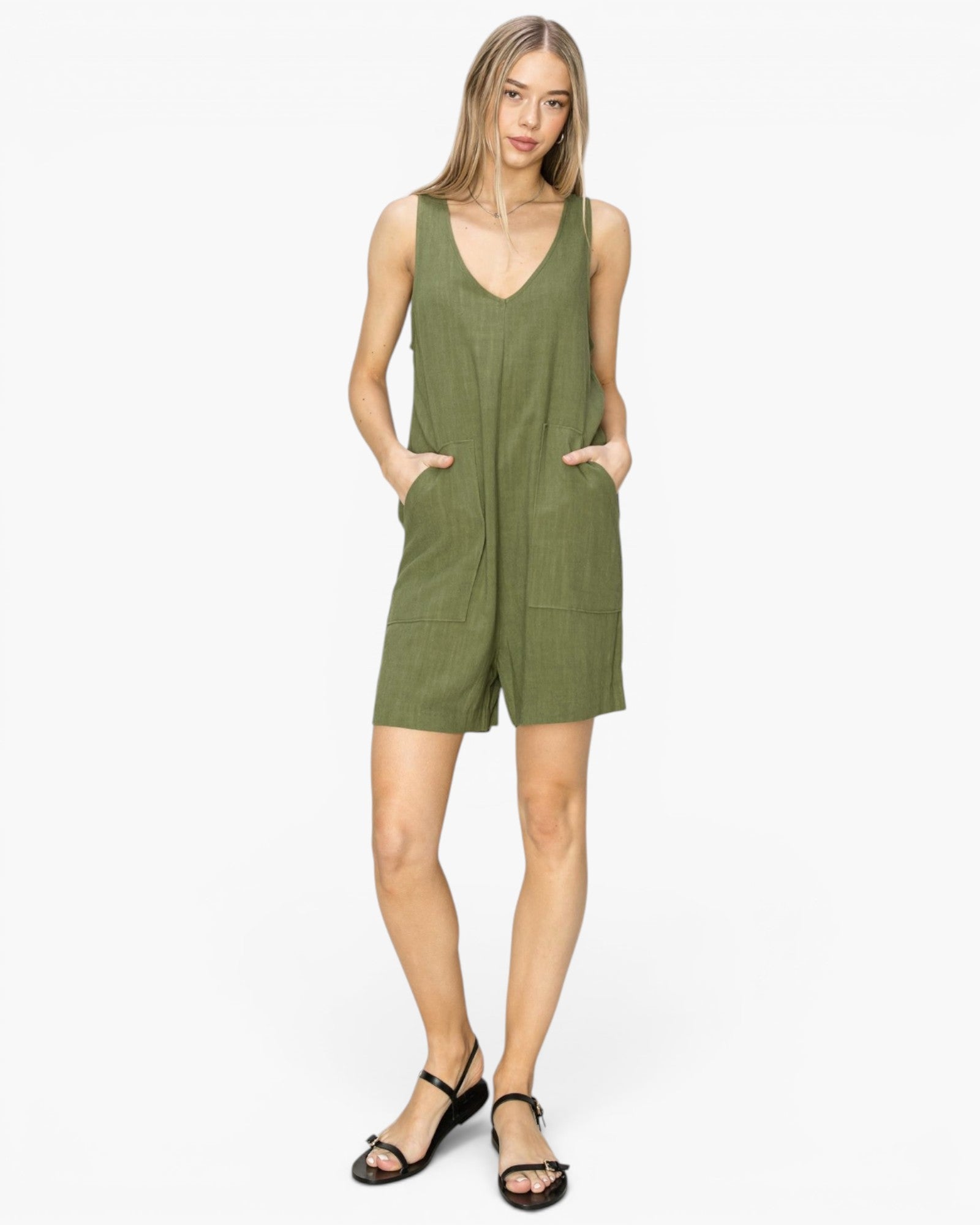 A woman modeling a sleeveless olive green romper with a V-neckline, front patch pockets, and short legs, paired with black strappy sandals against a plain white studio background.