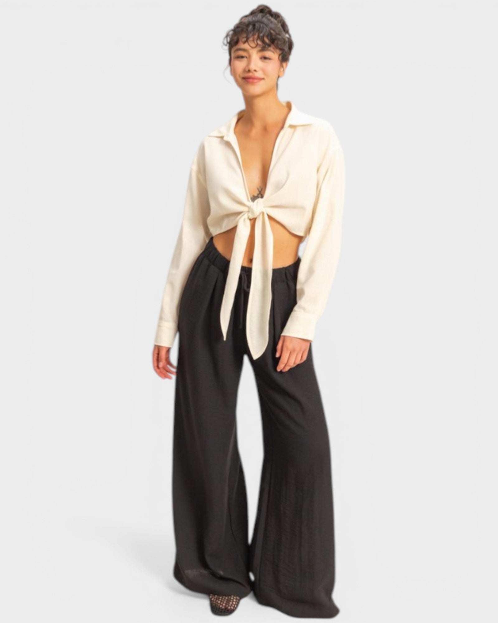 Woman wearing a cream blouse with a tie detail and black pants on a white background