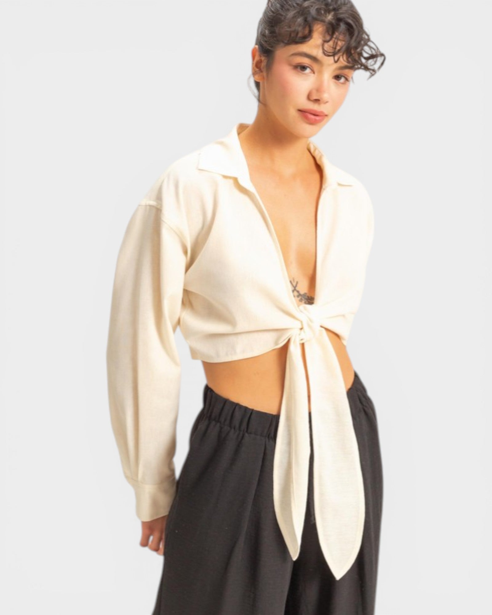 Woman wearing a cream-colored open blouse with a tie detail over a black top, on a light gray background.