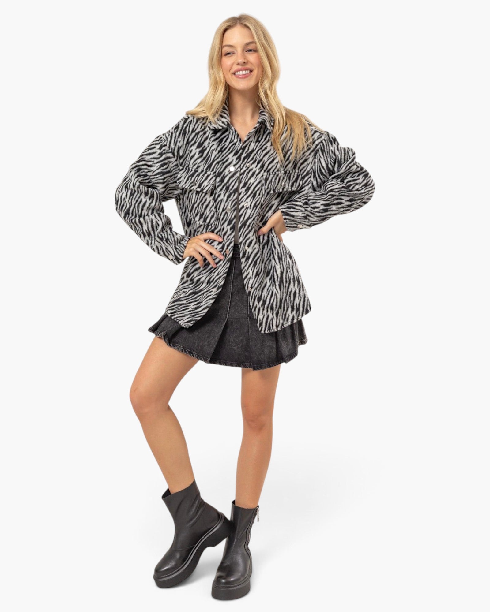 A woman modeling a black and white zebra-print oversized button-up shirt jacket, styled with a black pleated mini skirt and black leather ankle boots, against a plain white studio background.
