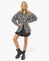 A woman modeling a black and white zebra-print oversized button-up shirt jacket, styled with a black pleated mini skirt and black leather ankle boots, against a plain white studio background.