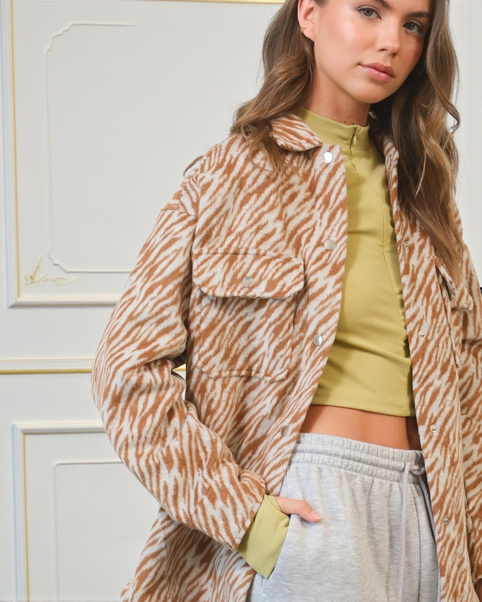 Woman wearing a patterned jacket over a green top and gray pants against a white wall.