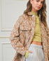 Woman wearing a patterned jacket over a green top and gray pants against a white wall.