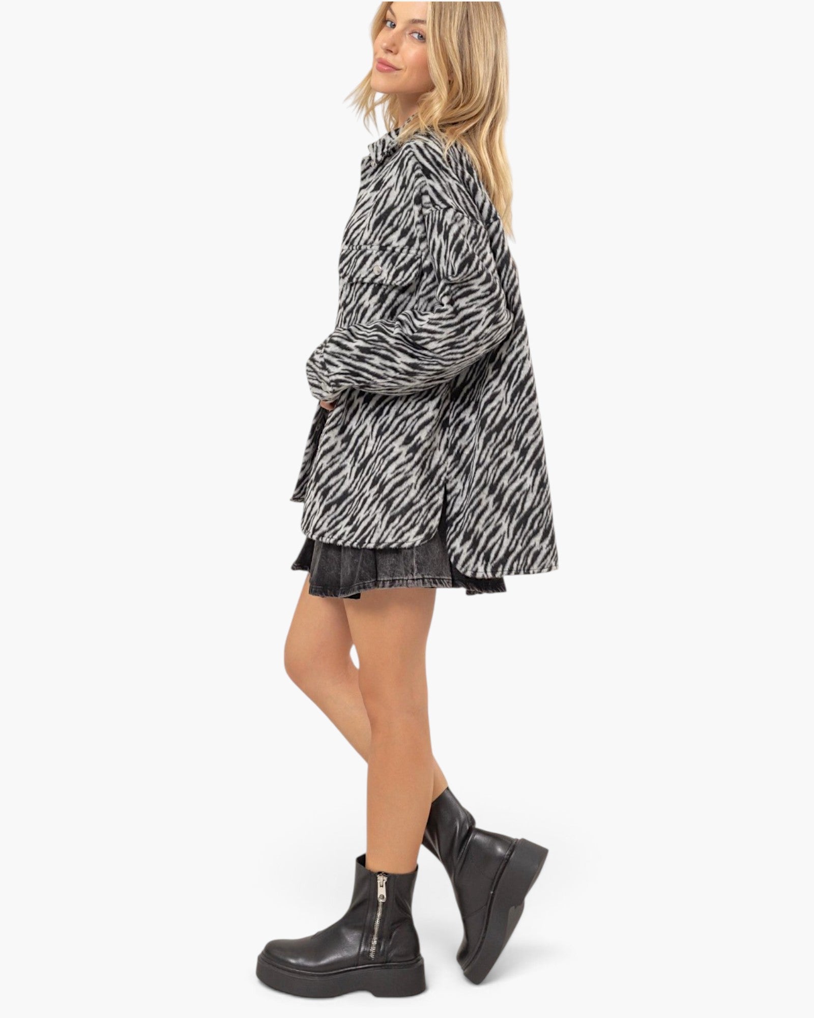 A woman modeling a black and white zebra-print oversized shirt dress with a ruffled hem, paired with black denim shorts and black platform ankle boots, against a plain white background.