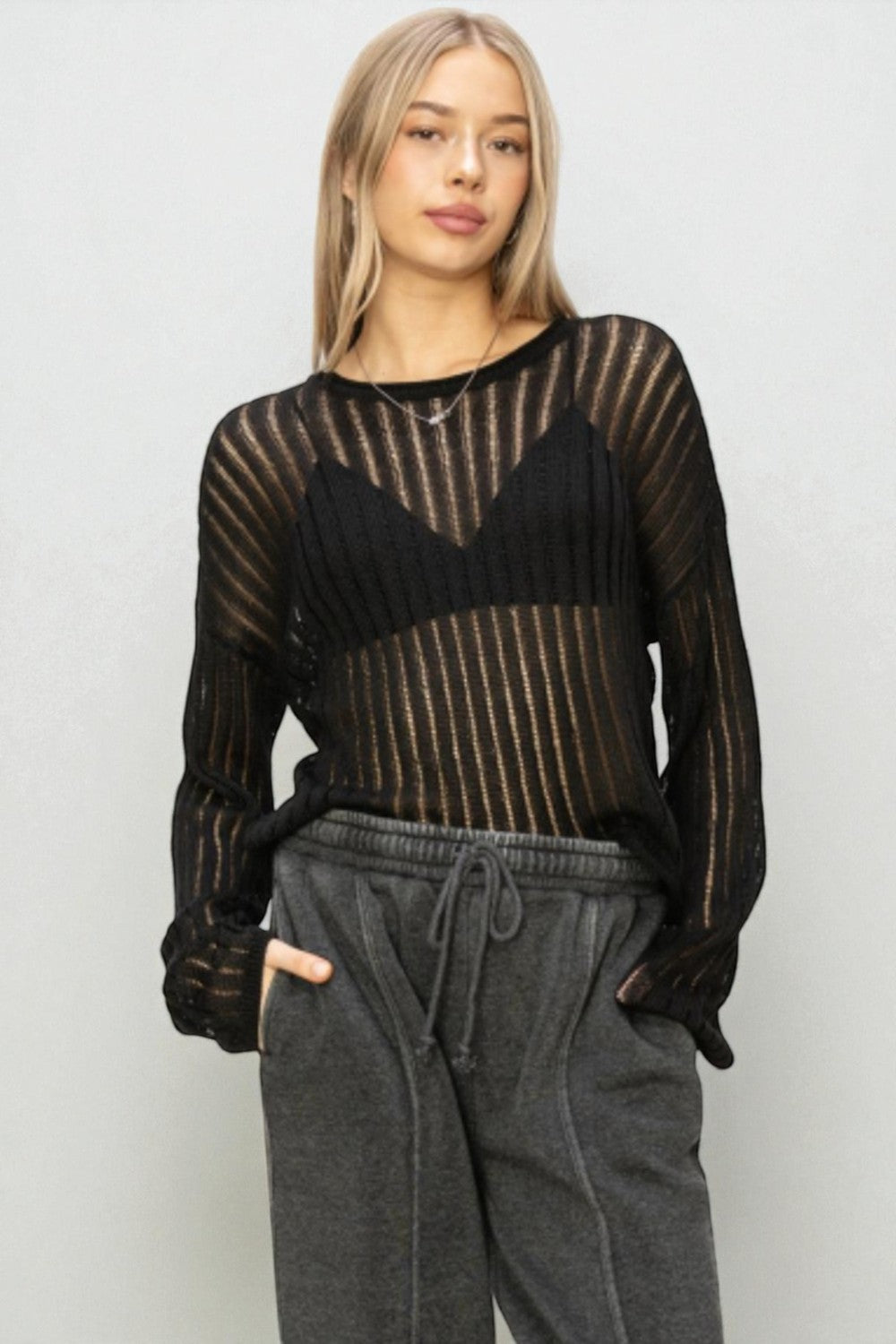 Woman wearing a black sheer long-sleeve top and gray sweatpants against a white background