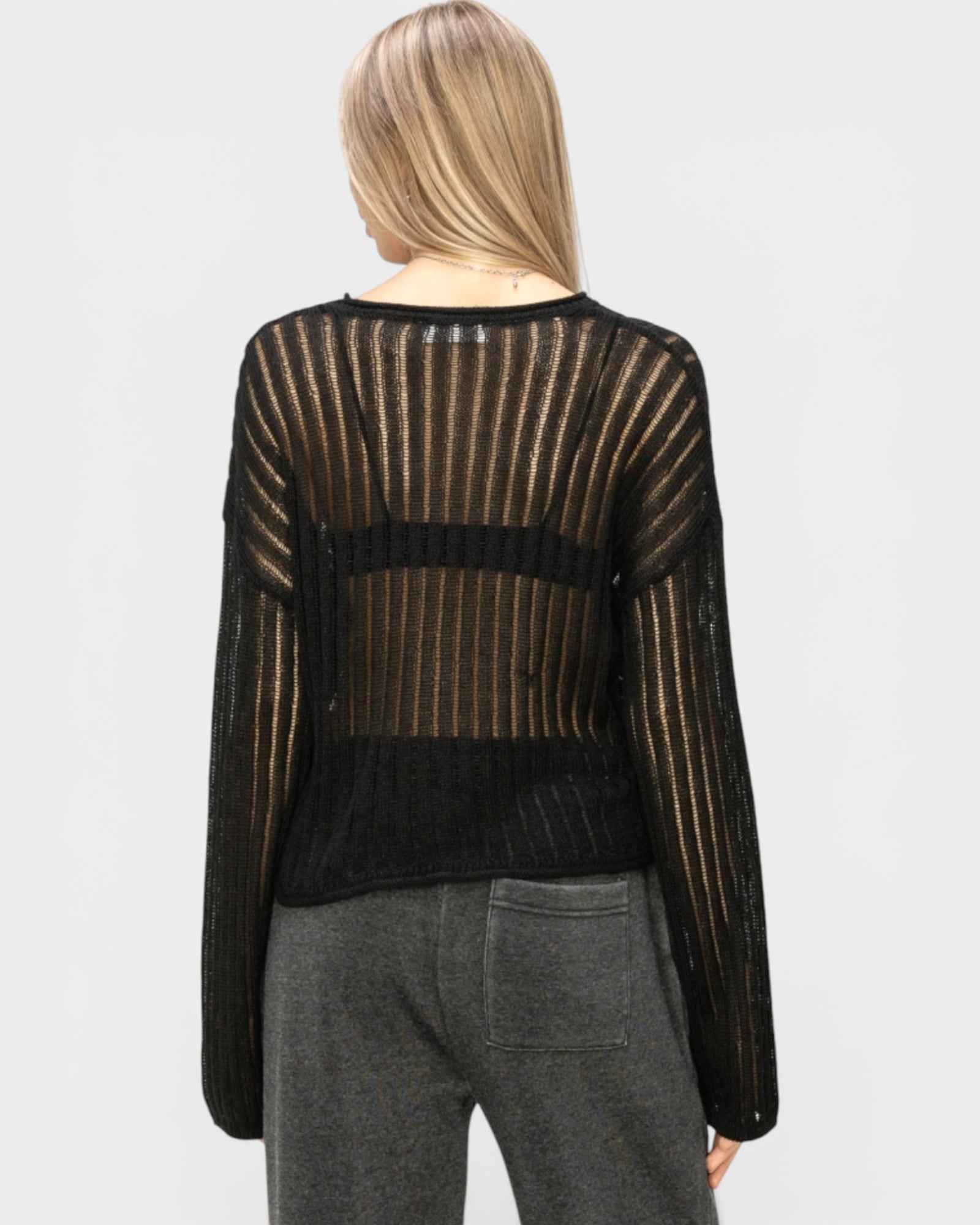 Person wearing a black sheer sweater over a black top with gray pants on a white background