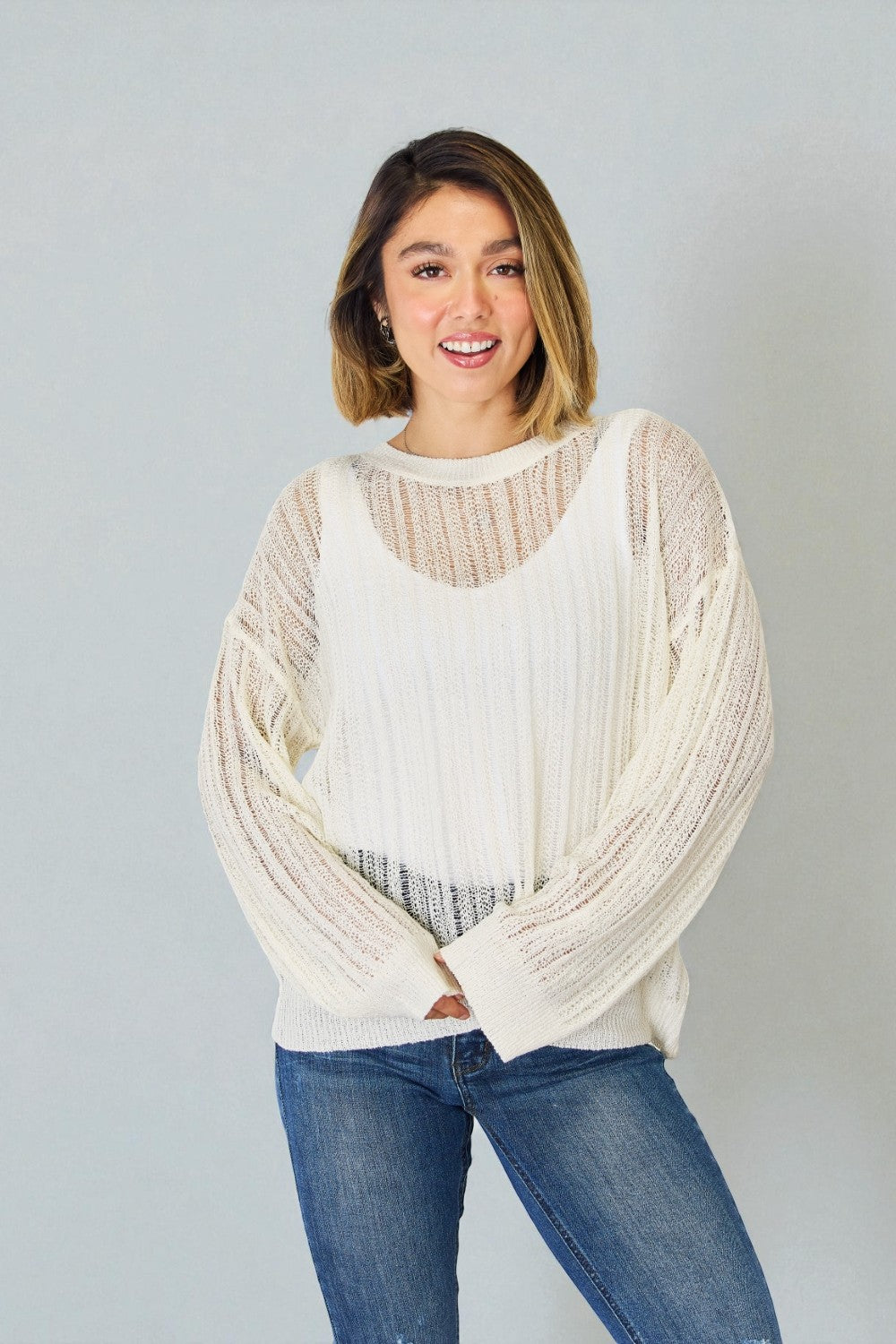Woman wearing a cream chunky knit sweater over a white shirt with blue jeans on a plain background