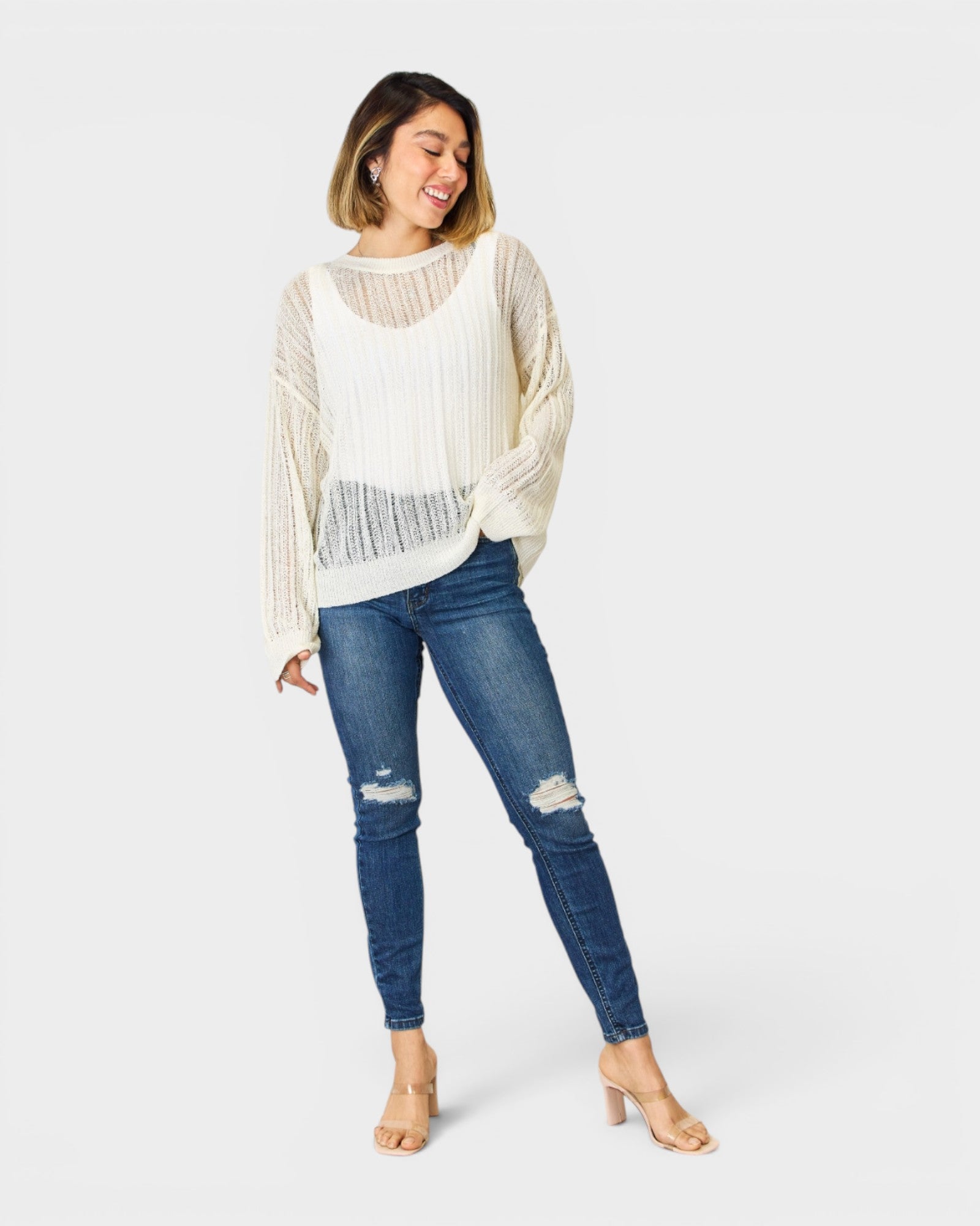 Woman wearing a white knit cardigan, white top, and blue jeans on a white background