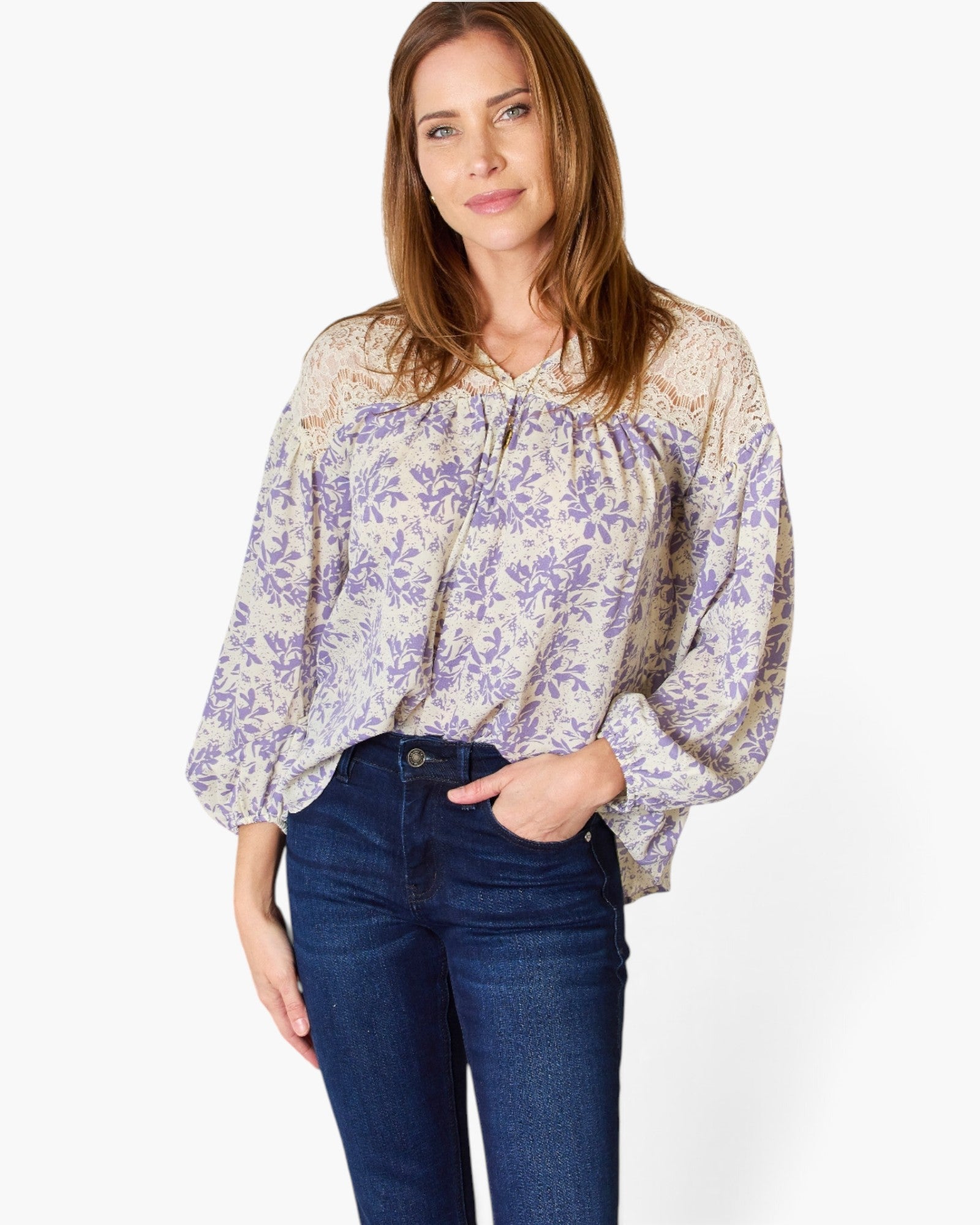 A woman wearing a long-sleeve blouse with a purple floral pattern on a cream background, paired with dark blue jeans, against a plain white studio background.