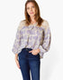A woman wearing a long-sleeve blouse with a purple floral pattern on a cream background, paired with dark blue jeans, against a plain white studio background.