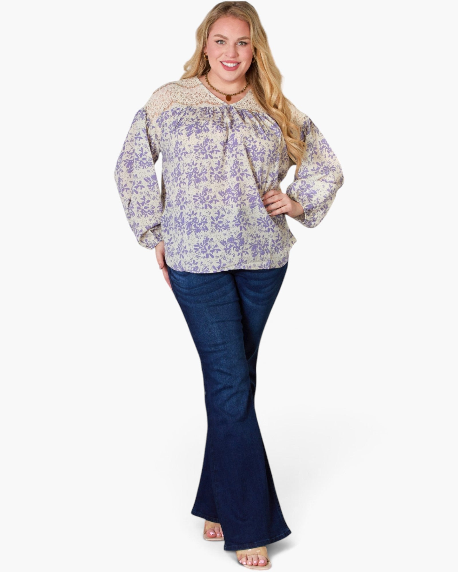 A woman modeling a long-sleeve blouse with a beige and lavender floral pattern and dark blue flared jeans, standing against a plain white studio background.